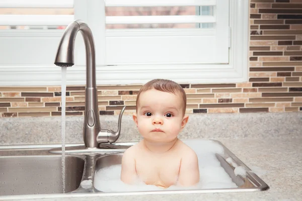 Portrait of cute Caucasian funny baby girl boy with dark black eyes sitting in big kitchen sink with water and foam  near window looking in camera, lifestyle everyday concept