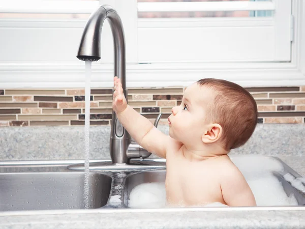 Portrait of cute Caucasian funny baby girl boy with dark black eyes sitting in big kitchen sink with water and foam  near window looking away, lifestyle everyday concept