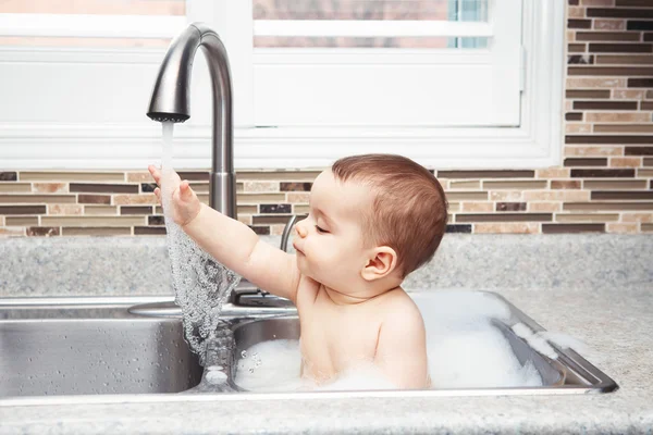 Portrait of cute Caucasian funny baby girl boy with dark black eyes sitting in big kitchen sink with water and foam  near window looking away, lifestyle everyday concept