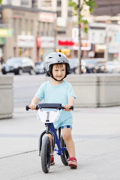 Portrait of smiling little boy toddler riding a balance bike bicycle in helmet on the road outside outdoors on spring summer day, seasonal child activity concept, healthy childhood lifestyle