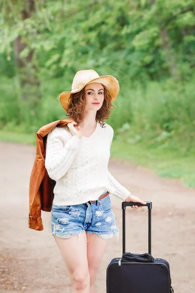 Portrait of beautiful Caucasian young girl woman in white sweater, blue denim shorts, straw hat, standing with travel bag on the wild country road in forest looking in camera, wanderlust adventure vacation
