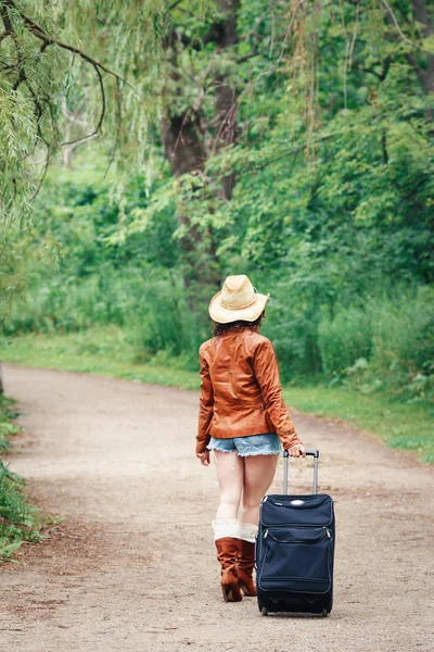 Portrait of Caucasian girl woman in leather jacket, blue denim shorts, straw hat, standing walking on country road wild forest with travel bag with her back to camera, wanderlust adventure vacation