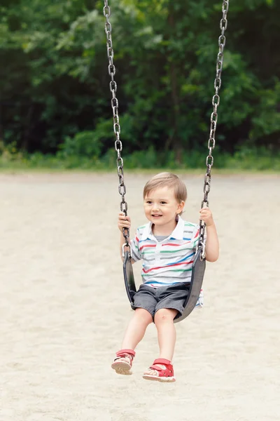Portrait of happy smiling little boy toddler in tshirt and jeans shorts on swing on backyard playground outside on summer day, happy childhood lifestyle concept