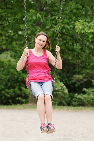 Portrait of happy smiling young middle age woman girl in red tshirt and jeans shorts on swing on backyard playground outside on summer day, lifestyle