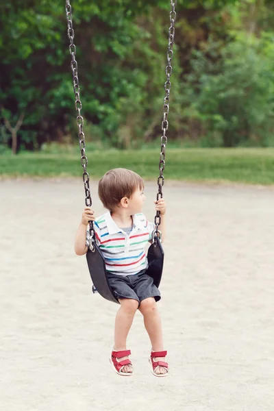 Portrait of cute little small boy toddler looking away, wearing tshirt and jeans shorts on swing on backyard playground outside on summer day, happy childhood lifestyle