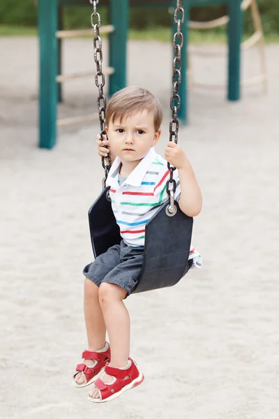 Portrait of sad upset worried little small boy toddler in tshirt and jeans shorts on swing on backyard playground outside on summer day, childhood lifestyle concept
