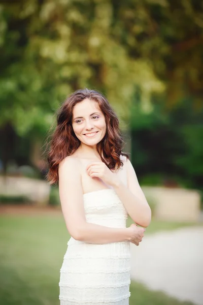 Portrait of beautiful smiling white Caucasian girl woman with long dark red brown hair and hazel eyes, in white summer  dress, standing in summer park outside, looking away, copyspace for text