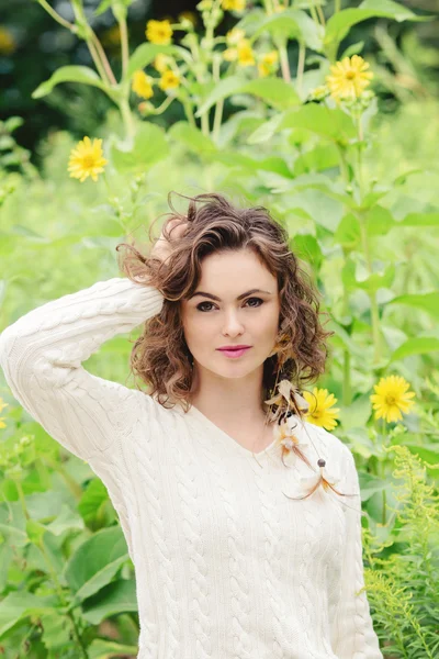 Portrait of beautiful smiling young white Caucasian girl woman touching her dark brown hair, in white sweater, standing in summer park field outside in green grass and yellow flowers, looking in camera
