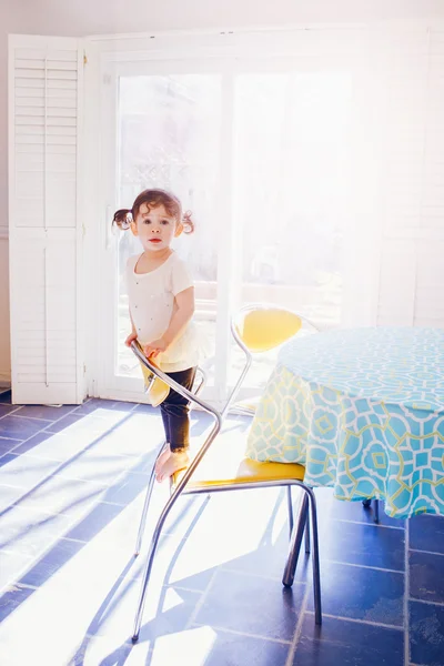 Portrait of adorable toddler girl in white dress with curly hair pig-tails standing on chair in kitchen looking in camera early morning, sunlight beams from window, touching everyday lifestyle moment
