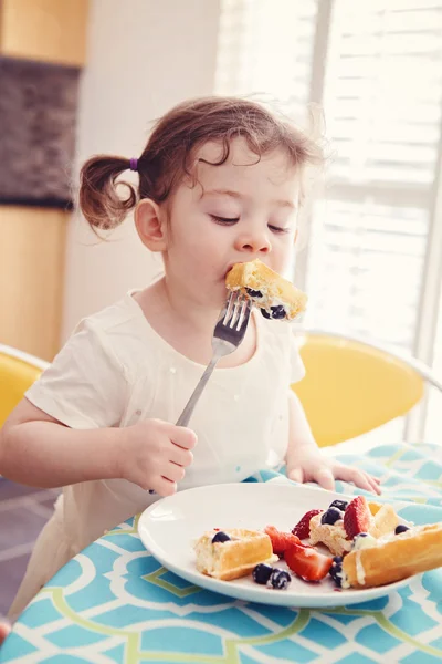 Portrait of one happy white Caucasian kid girl toddler with pig-tails in white dress eating breakfast waffles fruits with fork in sunny kitchen early morning