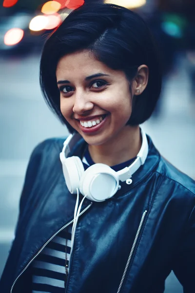 Portrait of beautiful Hispanic latino girl woman shor black hair in leather jacket with headphones outside in evening night city street smiling laughing looking in camera, lifestyle portrait concept