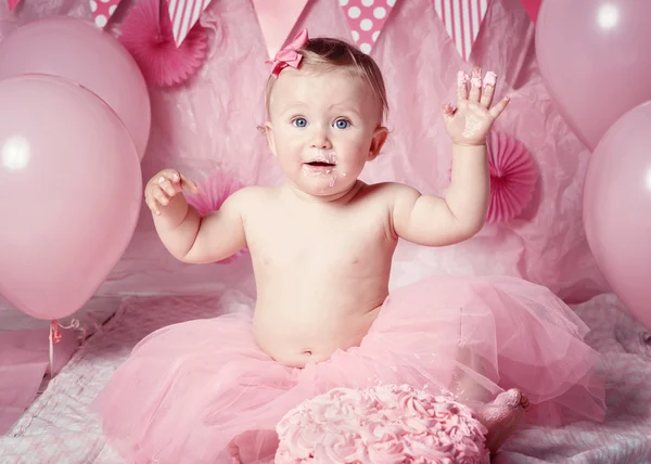 Portrait of cute adorable Caucasian baby girl with blue eyes in pink tutu skirt celebrating her first birthday with gourmet cake and balloons looking in camera, cake smash first year concept