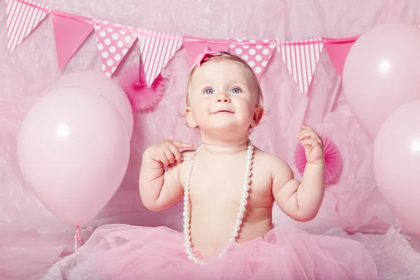 Portrait of cute adorable Caucasian baby girl with blue eyes in pink tutu skirt and pearls celebrating her first birthday with balloons looking away, cake smash first year concept
