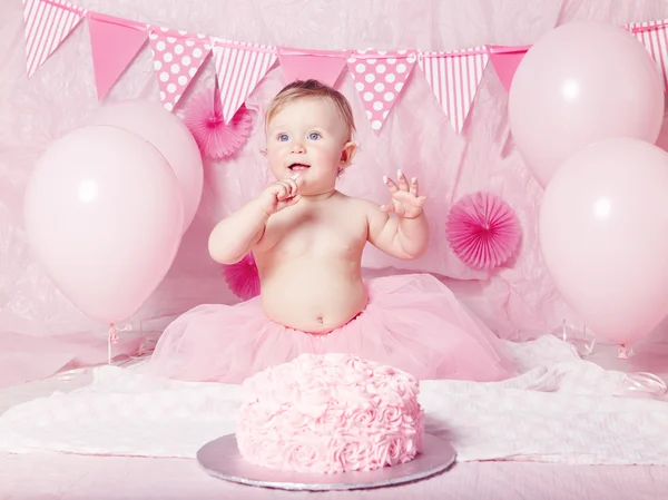 Portrait of cute adorable Caucasian baby girl with blue eyes in pink tutu skirt celebrating her first birthday with gourmet cake and balloons looking away, cake smash first year concept