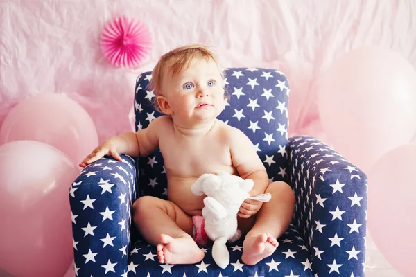 Portrait of cute adorable Caucasian baby girl with blue eyes sitting in blue children kids armchair with white stars celebrating her first birthday with balloons looking away