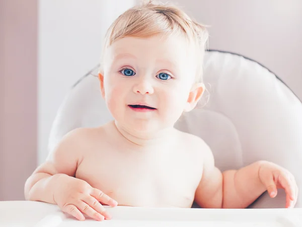Closeup portrait of cute adorable funny blonde Caucasian smiling laughing baby boy girl with blue eyes with emotional face expression sitting in high chair in kitchen looking away