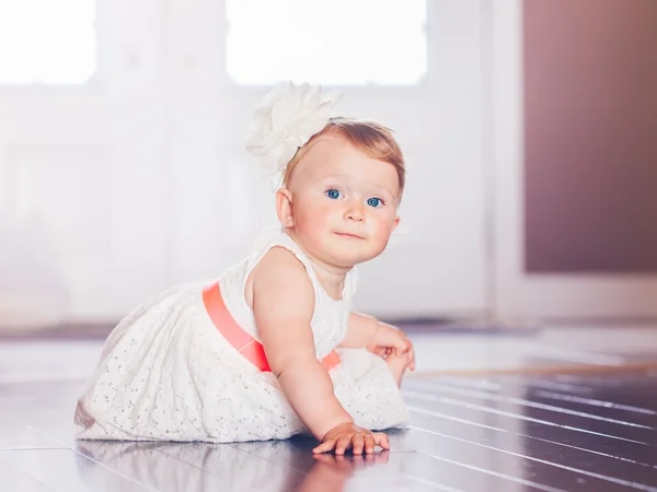 Portrait of cute adorable blonde Caucasian smiling baby child girl with blue eyes in white dress with red bow sitting on floor indoors looking in camera dreaming, fairy tale sun light from above behind