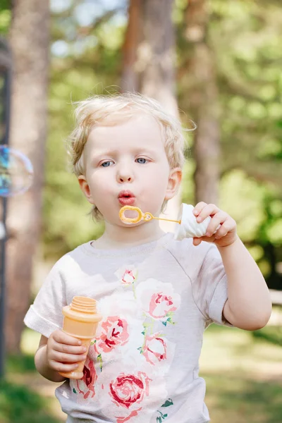 Portrait of cute funny little blond Caucasian child girl toddler standing in the green forest field meadow blowing soap bubbles, bright summer day, summer fun