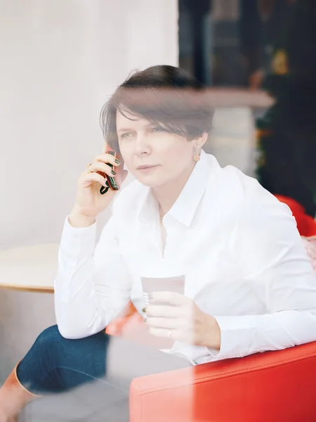 Closeup portrait of middle age Caucasian white business woman sitting in cafe restaurant with cup of coffee talking over on phone, shot through window glass with reflections, lifestyle candid concept