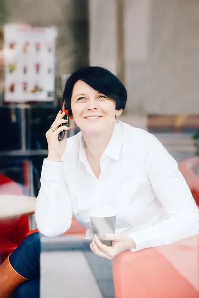 Closeup portrait of middle age Caucasian white business woman sitting in cafe restaurant with cup of coffee talking over on phone, shot through window glass with reflections, lifestyle candid concept