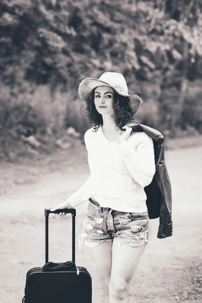 Black and white portrait of beautiful Caucasian young girl woman in white sweater, shorts, straw hat, standing with travel bag on the wild country road in forest looking in camera, wanderlust adventure vacation