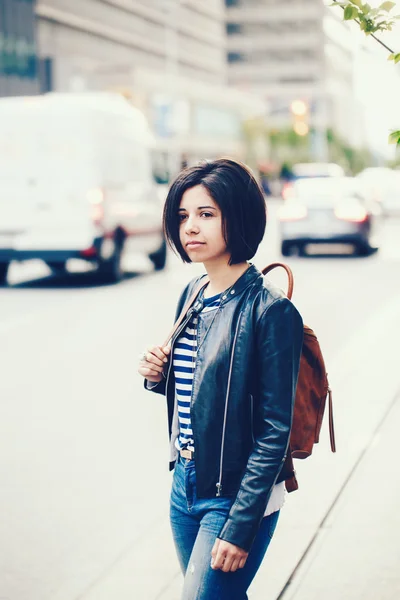 Portrait of beautiful young Caucasian latino girl woman with dark brown eyes and short dark hair in blue jeans, leather biker jacket with backpack standing in street outside, urban city view background