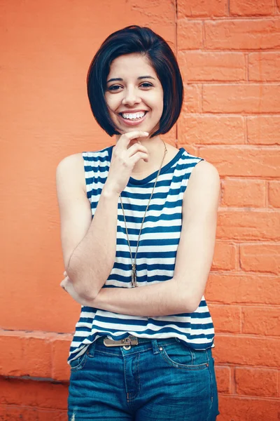 Portrait of beautiful smiling young hipster latin hispanic girl woman with short hair bob, in blue jeans, striped tshirt, leaning on red brick wall in city looking in camera, toned with Instagram filters