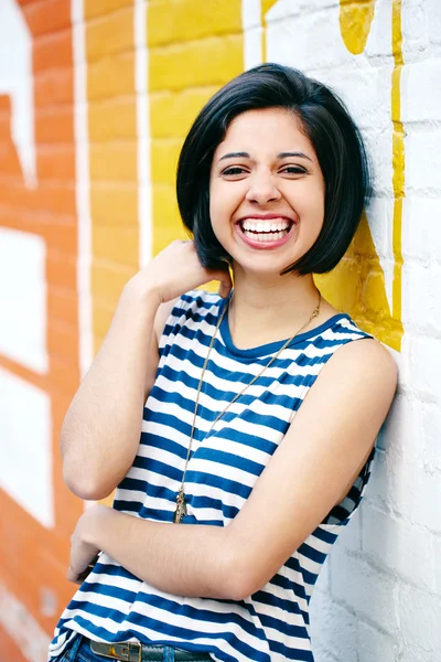 Portrait of beautiful laughing young hipster brunette latin hispanic girl woman with short hair bob, in blue jeans, striped tshirt, leaning on brick wall in city looking in camera