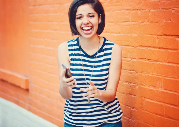 Portrait of beautiful young latin hispanic girl woman in striped tshirt and blue jeans with dark short long bob haircut outside near red brick wall in city making selfie photo with cell phone