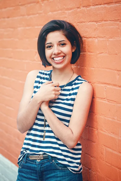 Portrait of beautiful smiling young hipster latin hispanic girl woman with short hair bob, in blue jeans, striped tshirt, leaning on red brick wall in city looking in camera, toned with Instagram filters