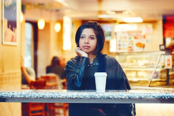 Candid portrait of beautiful young hipster latin hispanic girl woman with short hair bob, in grey cape poncho jacket, sitting in cafe restaurant with coffee looking away, shot through window glass