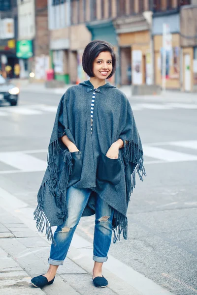 Portrait of beautiful smiling young hipster latin hispanic girl woman with short hair bob, in torn jeans, grey blue poncho cape, standing outside in city, looking in camera, street fashion concept