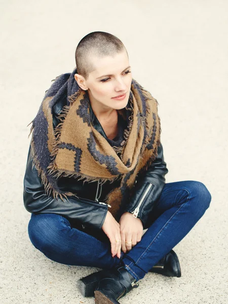 Portrait of beautiful Caucasian white young bald girl woman sitting on ground outside with shaved hair head in jacket, scarf shawl, jeans, looking away, toned with Instagram filters in yellow color