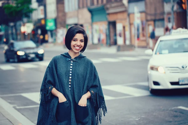 Portrait of beautiful smiling young hipster latin hispanic girl woman with short hair bob, in torn jeans, grey blue poncho cape, standing outside in city street, looking in camera, street fashion concept