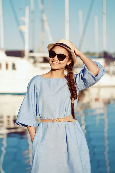 Portrait of smiling white Caucasian brunette woman with tanned skin in blue dress hat, by seashore lakeshore with blurry yachts boats on background on water, sunset on summer day, lifestyle concept