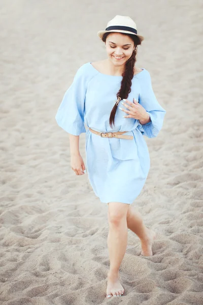 Portrait of smiling laughing white Caucasian brunette woman with tanned skin in blue dress and straw hat running on sand beach, sunset on summer day, lifestyle concept