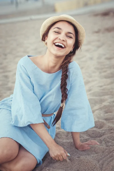 Portrait of smiling laughing white Caucasian brunette woman with tanned skin in blue dress and straw hat sitting on sand beach, sunset on summer day, lifestyle concept