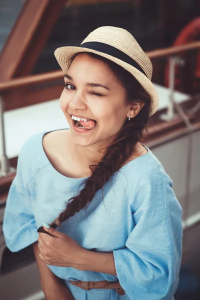 Closeup portrait of funny beautiful smiling white Caucasian brunette girl winking showing tongue, in blue dress and straw hat, by yacht boat, navy maritime retro vintage style concept