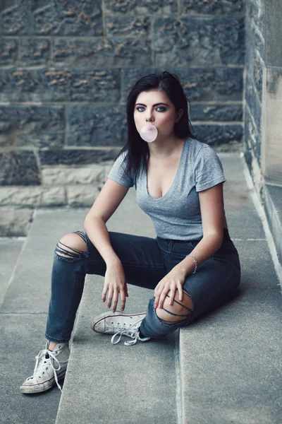 Portrait of white Caucasian beautiful young brunette girl with light blue green eyes in ripped jeans, t-shirt, keds. Teenager blowing bubble gum looking in camera. Toned with Instagram filters, lifestyle concept.
