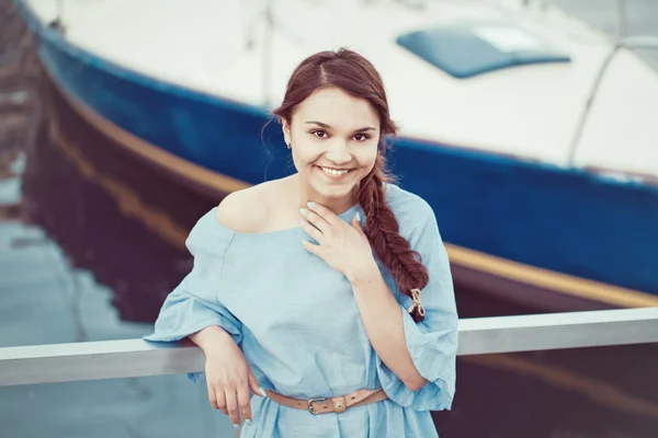 Portrait of white Caucasian brunette woman with tanned skin in blue dress by seashore lakeshore with yachts boats on background on water, lifestyle concept