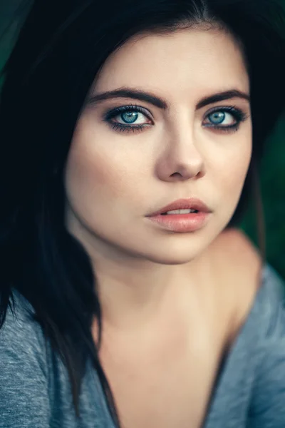 Closeup portrait of beautiful young Caucasian woman with black hair, blue eyes, looking in camera, sitting outdoors, toned with filters, natural beauty youth look