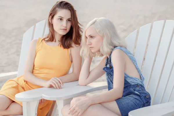 Portrait of two pensive sad white Caucasian unformal young girls hipster students friends outside on beach summer day sitting on chairs silent, best friends forever, selective focus