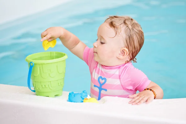 Portrait of white Caucasian baby girl playing with toys in water on swimming pool nosing inside, looking away, training to swim, healthy active lifestyle