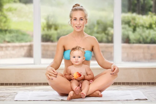 Group portrait of white Caucasian mother and baby daughter doing physical fitness exercises yoga together sitting in lotus pose on floor in swimming pool looking in camera, eating apple