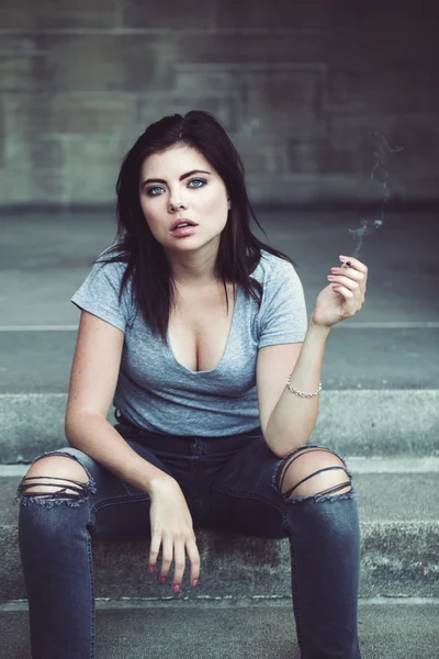 Portrait of white Caucasian beautiful young brunette woman teenager with blue green eyes in ripped jeans, t-shirt holding smoking cigarette looking in camera, toned with filters, lifestyle concept