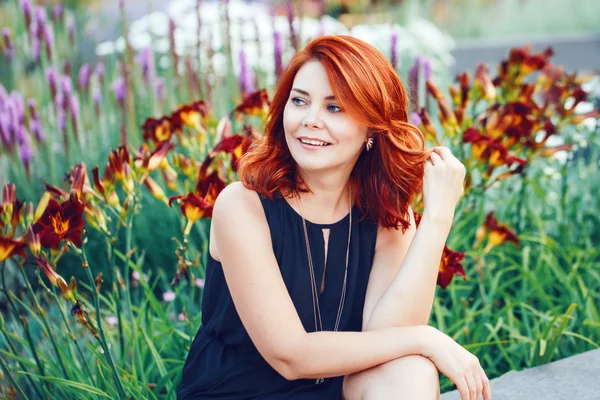 Closeup portrait of smiling middle aged white caucasian woman with waved curly red hair in black dress looking away outside in park garden among flowers, beauty fashion lifestyle concept