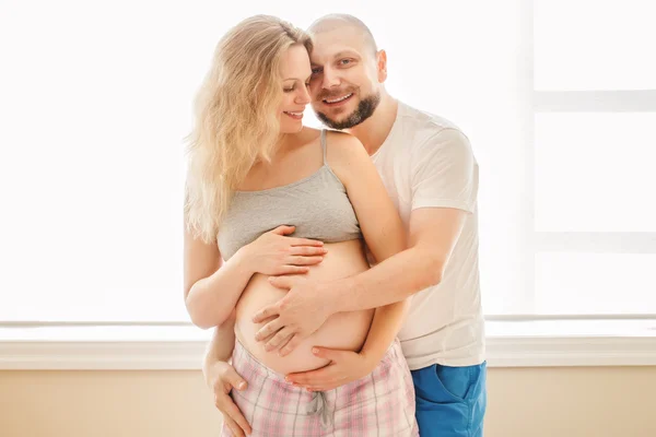 Portrait of smiling laughing white Caucasian young middle age couple, pregnant woman with husband in room hugging cuddling, lifestyle maternity concept
