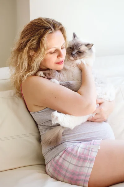 Portrait of young white Caucasian blonde pregnant woman with long hair in tshirt and shorts relaxing at home on  couch holding cat, healthy maternity lifestyle concept