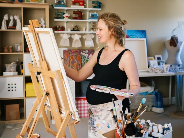Portrait of smiling white Caucasian young pregnant woman drawing painting standing at easel in home studio, art mental therapy, healthy happy lifestyle concept