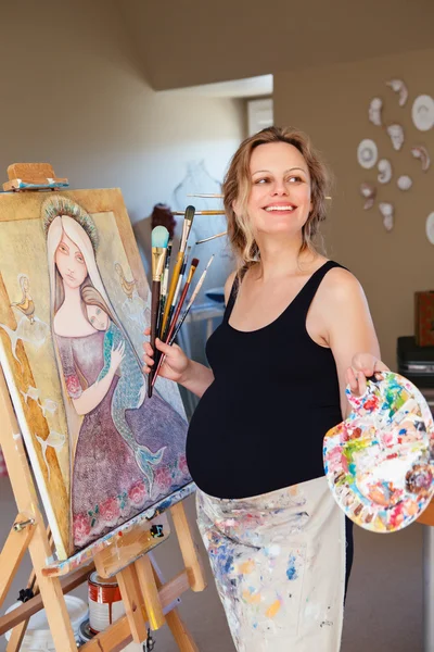 Portrait of smiling white Caucasian young pregnant woman drawing painting standing at easel in home studio, art mental therapy, healthy happy lifestyle concept